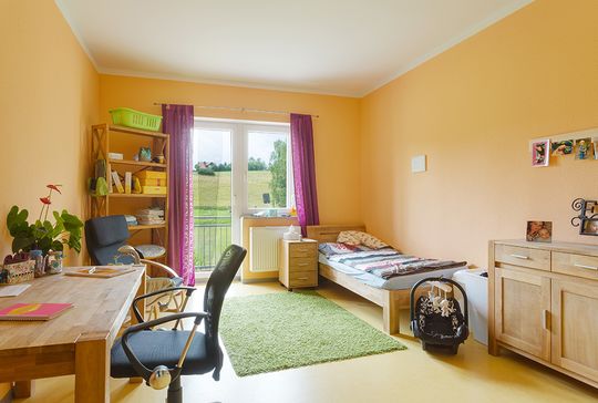 Blick durch die Tür in ein gemütlichen Mutter-Kind-Zimmer im Suchthilfezentrum Wolfersdorf. Tisch, Schrank und ein großer Schreibtisch sind aus hellem Holz. Violett gemusterte Stores vor dem Balkonfenster, ein grüner flauschiger Teppich und ein warmes Gel