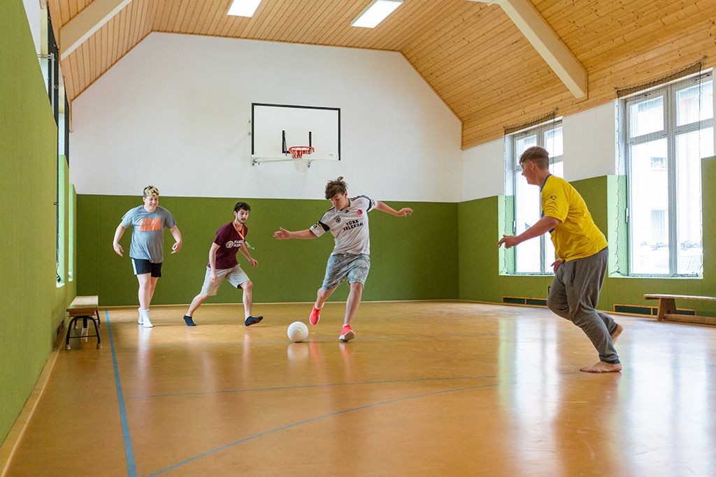 Vier Jungs in kurzen Hosen spielen in der kleinen Turnhalle des Jugendhilfezentrums Wolfersdorf Fußball. Der Junge in der Mitte des Bildes holt gerade mit seinem Bein aus, um den Ball seinem Gegenüber zuzuspielen.