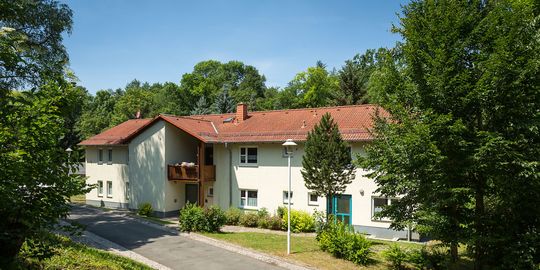 Zwischen grünen Bäumen sieht man ein modernes zweistöckiges Haus mit Holzbalkonen, in dem Jugendliche im Jugendhilfezentrum Bad Köstritz wohnen.