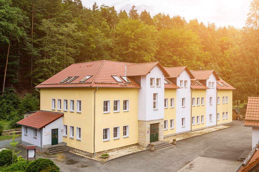 Blick auf das gelbe dreistöckige Gebäude des Jugendhilfezentrums Wolfersdorf. Hinter dem Gebäude sieht man Mischwald, hinter dem die Sonne untergeht.