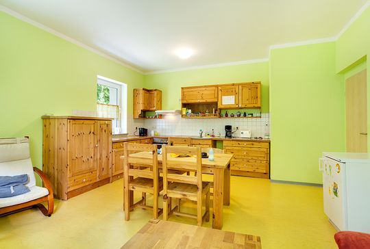 Blick in die gemütliche Küche der Bewohnerinnen im Suchthilfezentrum Wolfersdorf. Ein Holztisch mit 4 Stühlen steht in der Mitte des großen Raumes. Ein Schwingsessel mit hellem Bezug, eine Küchenzeile aus Holz und eine Waschmaschine  sind im Raum verteilt