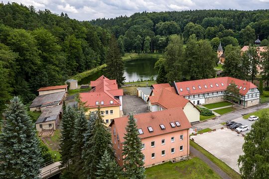 Luftansicht auf das große Areal des Jugendhilfezentrums Woltersdorf. Man sieht die heimeigene Schule und die Wohngruppenhäuser, welche sich idyllisch mitten im Wald an einem See befinden.