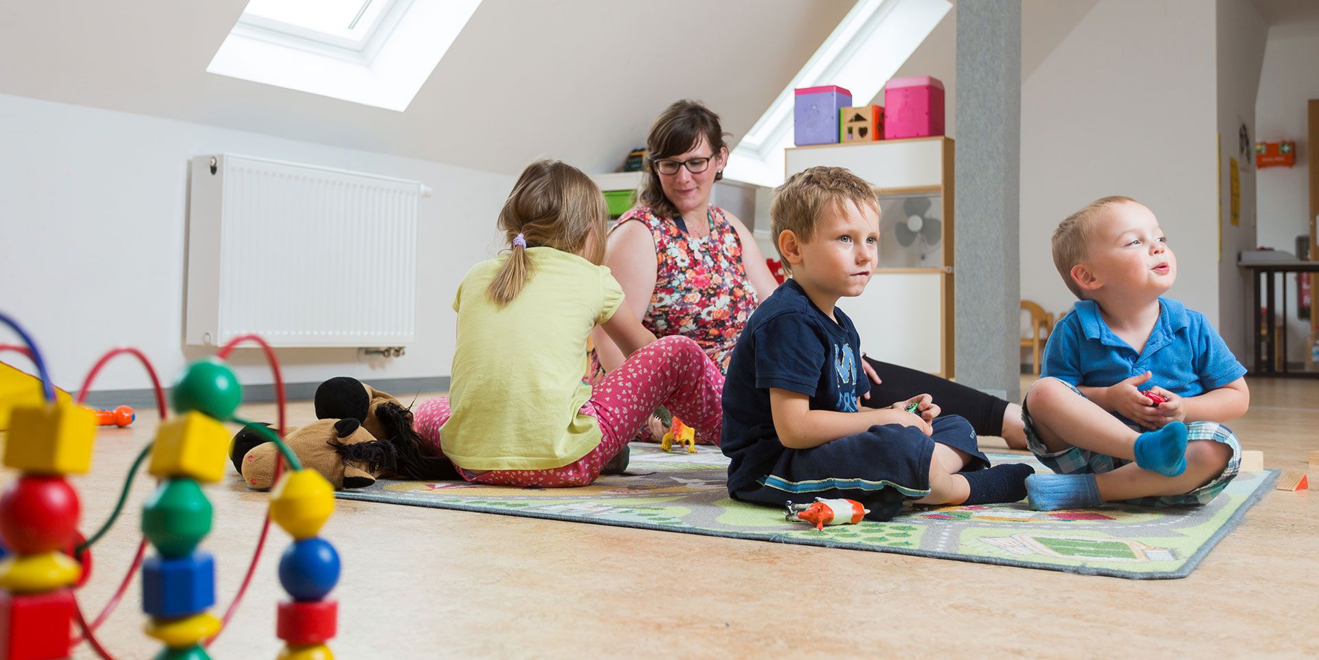 Junge Erzieherin mit Brille spielt mit Kindern auf buntem Spielteppich im Straßendesign