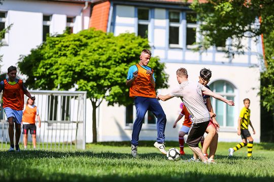 Eine bunte Gruppe Jugendlicher kickt auf dem Rasenplatz vor der Förderschule Fußball.