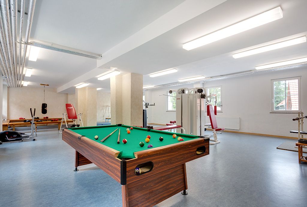 Blick in den lichtdurchfluteten Kraftraum. Im Vordergrund des großen Raumes mit grauem Fußboden sieht man einen modernen Billard-Tisch, im Hintergrund  befinden sich die unterschiedlichsten Kraftgeräte.